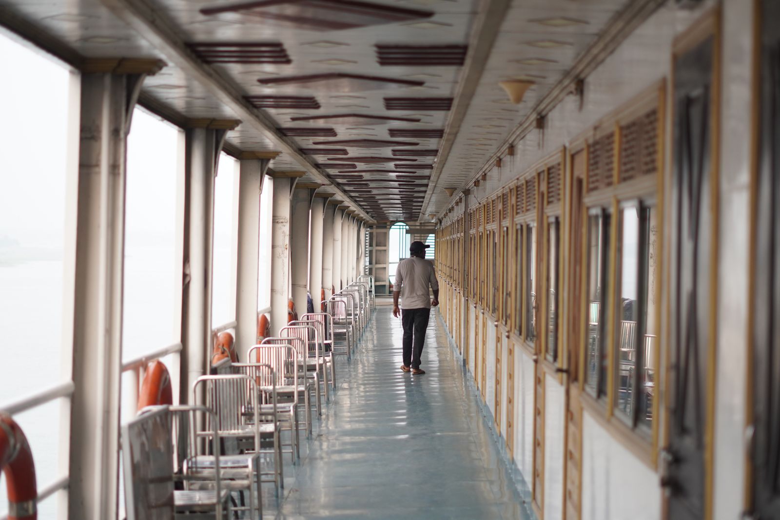A passenger walking along a covered promenade on a ship, lined with windows and deck chairs
