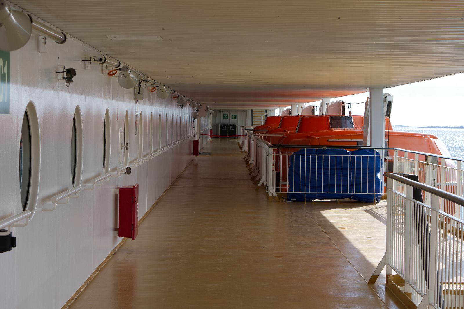 A covered promenade deck with portholes on one side and orange lifeboats on the other