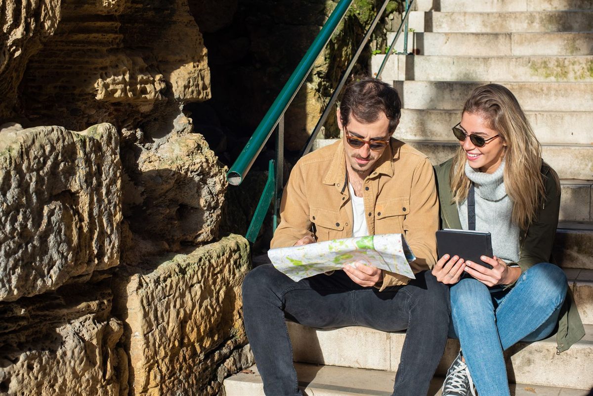 A couple on stone steps planning a trip with a paper map and a tablet